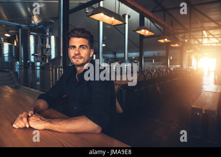 Portrait of handsome young man sitting at table with tanks in background at brewery. Caucasian man at beer tasting area. Stock Photo