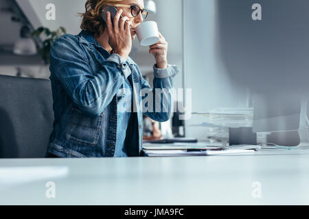 Female at work drinking coffee and talking on mobile phone. Young businesswoman at work making phone call and having coffee. Stock Photo