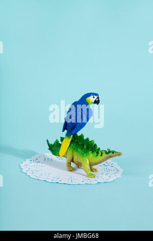 A dinosaur stegosaurus and a blue parrot on a white lace paper doily with a vibrant turquoise background. Minimal offbeat still life photography Stock Photo