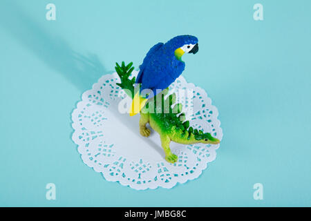 A dinosaur stegosaurus and a blue parrot on a white lace paper doily with a vibrant turquoise background. Minimal offbeat still life photography Stock Photo