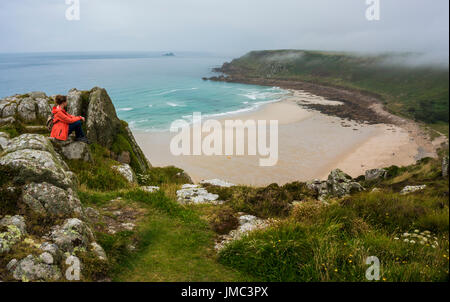 Young woman sitting by herself looking out at empty cove Gwynver Beach, Cornwall, England, UK Stock Photo