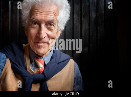 Senior man portrait. Close up, looking at camera. Smiling, well dressed, leaning against dark wall Stock Photo