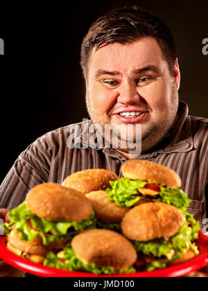 Fat man eating fast food hamberger and carries treat for friends on tray. Breakfast for overweight person. Poor food leads to obesity. Person regularl Stock Photo