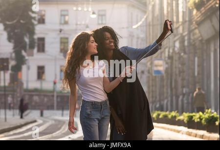 two young friends walking on the streets and making selfie. Black girl and her white friend. Stock Photo