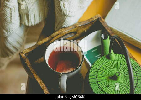 Off-white knitted sweater hanging over wooden chair, mug with red fruit tea, pot in tray by window, old book, hipster style, autumn, fall mood, toned, Stock Photo