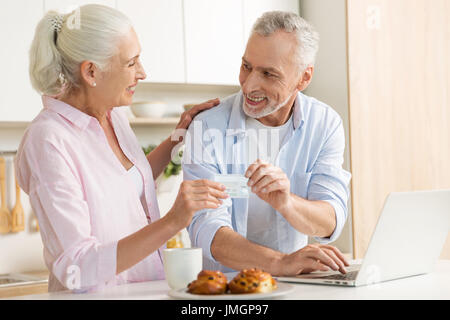 Picture of cheerful mature loving couple family standing at the kitchen using laptop computer holding credit card. Looking at each other. Stock Photo
