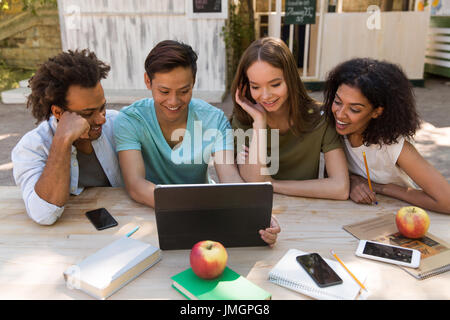 Photo of smiling young multiethnic friends students outdoors using tablet computer and talking with each other. Looking aside. Stock Photo