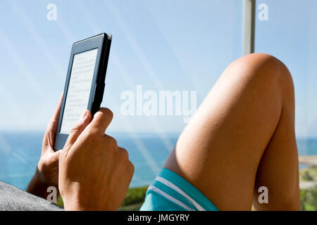 closeup of a young caucasian man wearing swimsuit reading in a tablet or e-reader in a balcony, with the ocean in the background Stock Photo