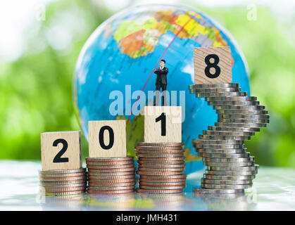 Tiny miniature businessman model is standing on a wooden numbers 2018 on growing stacks of coins with globe background using as growing business. Stock Photo