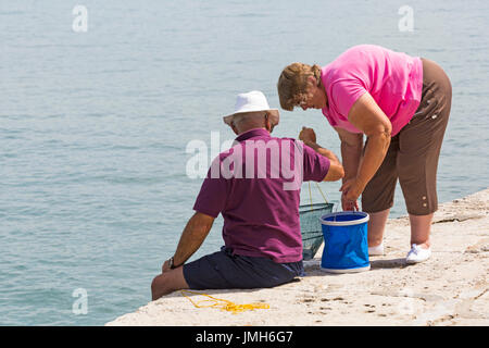 Man sitting on wall with basket net crabbing fishing with woman at Lyme Regis, Dorset in July Stock Photo
