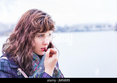 Young woman putting lipstick on in winter by river closeup portrait Stock Photo