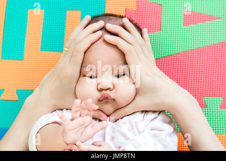 Hands of mother squeezing on face of her cute little baby on the eva foam in bedroom Stock Photo