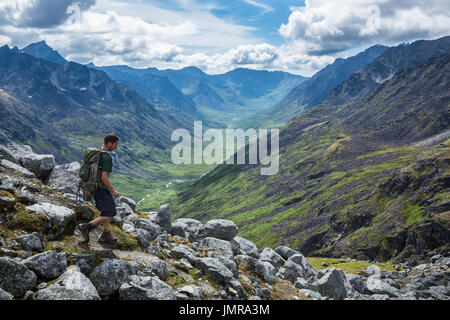 A man in his mid-20's hikes with a daypack down a steep trail overlooking the Little Susitna River in the Gold-Mint Trail Valley of the Hatcher Pass a Stock Photo