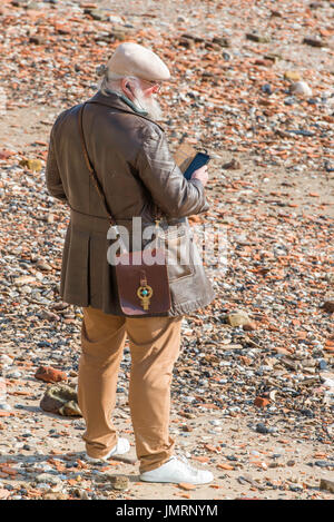 A mature man with a white beard and hair in a ponytail, standing on the banks of the Thames at low tide,  wearing headphones and holding a mobile. Stock Photo