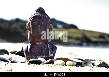 Meditating Buddha statue on sand in outdoor setting. In natural setting statue of Buddha at beach with pebble stones and sea waves and blue sky Stock Photo