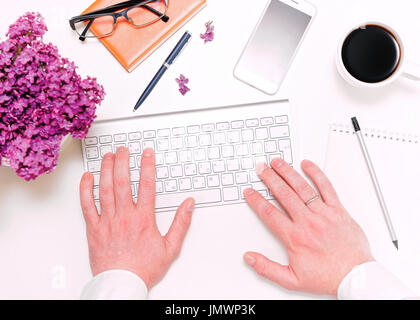 Man fingers working with computer keyboard and workplace with mobile phone, cup of coffee, notebook, pencil, pen, glasses and lilac flowers on the whi Stock Photo