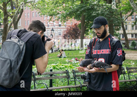 A tourist in Washington Square Park feeds pigeons from his hand while a friend commemorates it with a photo. New York City. Stock Photo