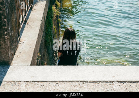 Young woman in a statement black t-shirt sits on steps by the water. Stock Photo