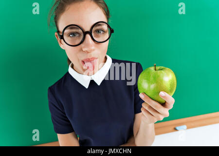 Emotional girl. Photo of sassy student shows tongue and holding an apple near blackboard, education concept Stock Photo