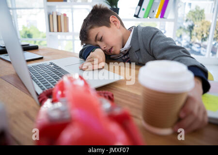 Boy as business executive sleeping while holding coffee cup in office Stock Photo
