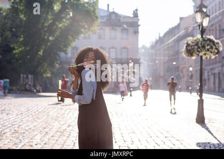 Happy attractive black woman blowwing bubbles on the street on a sunny day. Beautiful architecture around. Stock Photo