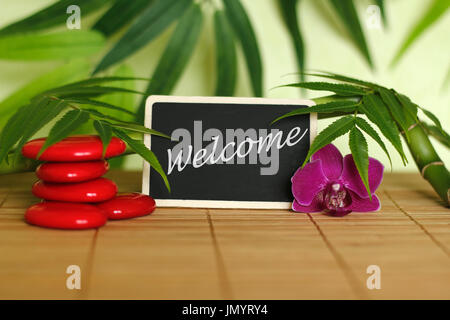 Red pebbles arranged in Zen lifestyle with an orchid, a lighted candle, a bamboo branch and foliage with the message welcome on the slate Stock Photo