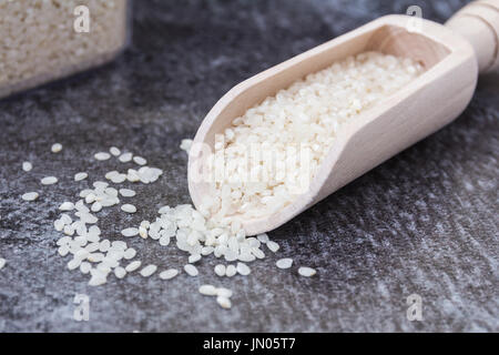 Wooden Scoop with Pudding Rice and Rice on a grey surface, with large container of Rice in the background Stock Photo