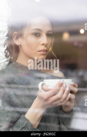 Thoughtful woman having coffee in cafe seen through window Stock Photo