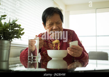 Senior woman using mobile phone while having noodles at home Stock Photo