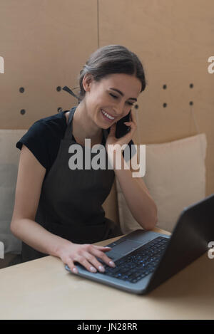 Owner talking on phone while using laptop at table in cafe Stock Photo