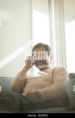 Smiling woman taking on phone while sitting on sofa at home seen through table glass Stock Photo