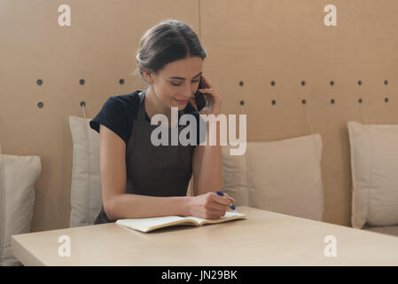 Owner talking on phone while writing on diary at table in cafe Stock Photo