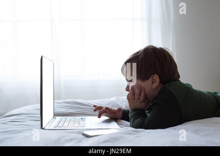 Side view of boy using laptop while lying on bed by window Stock Photo