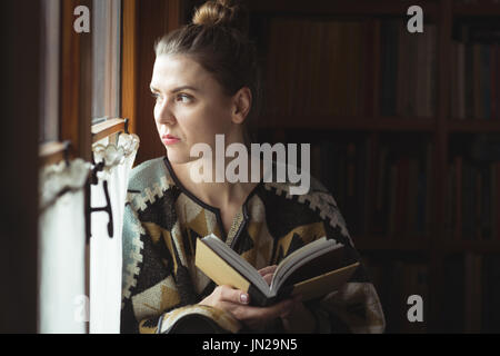 Thoughtful woman looking through window while reading book at home Stock Photo
