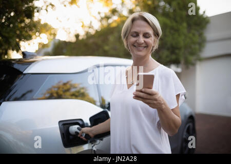 Smiling woman using mobile pone while charging electric car Stock Photo