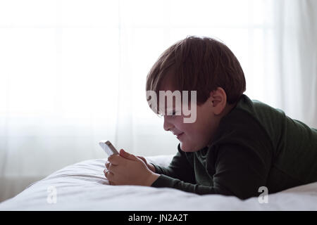 Side view of smiling boy using smart phone while lying on bed by window Stock Photo