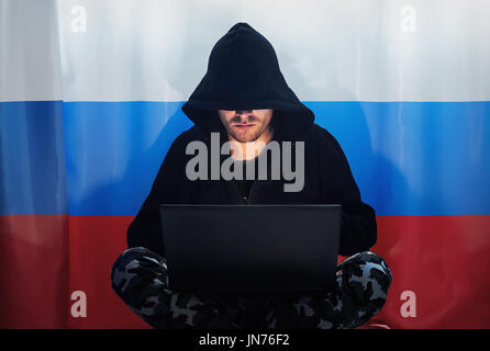 Hacker in a dark hoody sitting in front of a notebook on the background of the Russian flag. Stock Photo