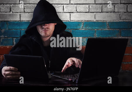 a hacker sitting at a Desk with a computer and a tablet in his hand and flag on background - Russia Stock Photo