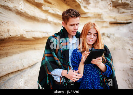 Beautiful woman and handsome man wrapped in a blanket. They are smiling and looking at the screen of a tablet on the background of a sand quarry. Stock Photo