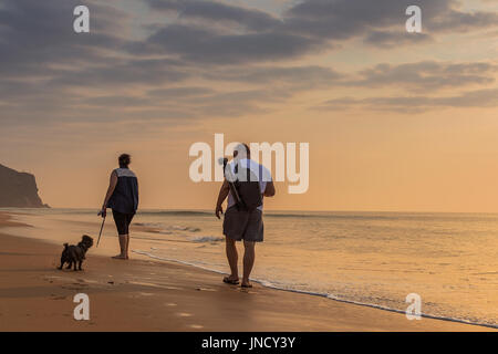 CABO LEDO/ANGOLA - 22 JULY 2017 - Couple walking on the beach with dog, sunset. Stock Photo
