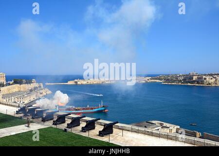 Soldiers firing the cannons for the Noon Gun in Upper Barrakka Gardens with views across the Grand Harbour towards Fort Rikasoli, Valletta, Malta, Eur Stock Photo