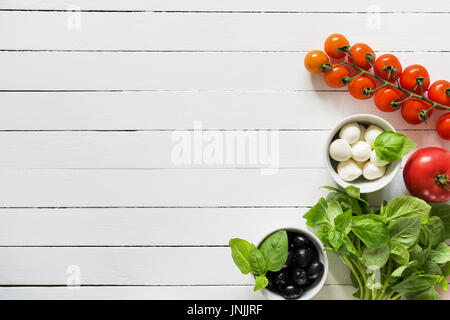 Fresh food ingredients for italian cuisine. Bunch of green basil, black olives, mozzarella cheese and tomatoes on white wooden background. Top view wi Stock Photo