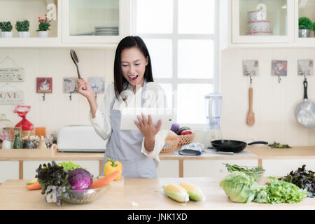 Cheerful Asian young woman is cooking in the kitchen with joy. She is standing and holding digital tablet of recipe. Asian woman is touching a wood sp Stock Photo