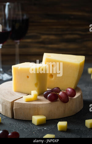 Swedish hard yellow cheese with holes chopped with red grapes on wooden slices and glasses with red wine on dark rustic background Stock Photo