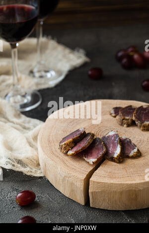 Slices of dried meat on wooden cut, red grapes and two glasses of red wine on dark rustic background