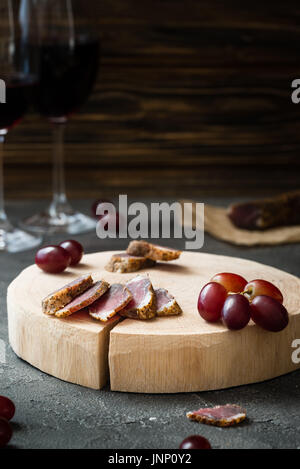 Slices of dried meat on wooden cut, red grapes and two glasses of red wine on dark rustic background