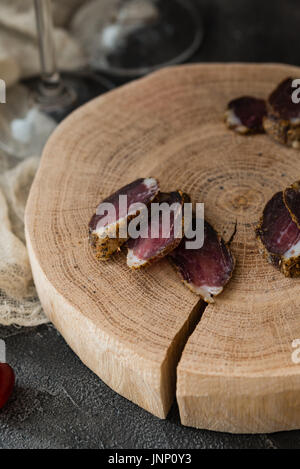 Slices of dried meat on wooden cut and two glasses legs on dark rustic background