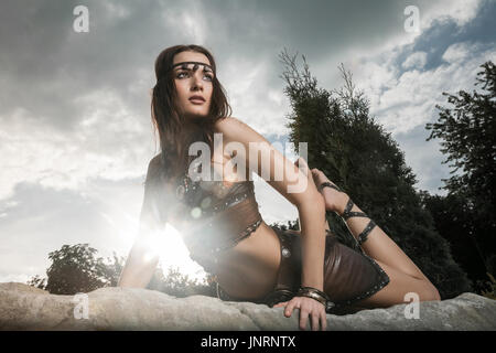 Young beautiful woman dressed in a leather wear looking like an amazon or warrior lying on a big stone. Dramatic beauty shot Stock Photo