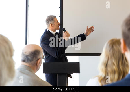 Business speaker with microphone pointing with hand at blank whiteboard in front of audience, conference, seminar concept Stock Photo