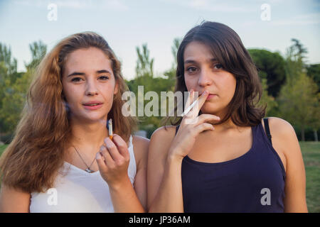 Two girls friends smoking cigarettes in the park on a cloudy day. They are of celebration and are happy. Stock Photo